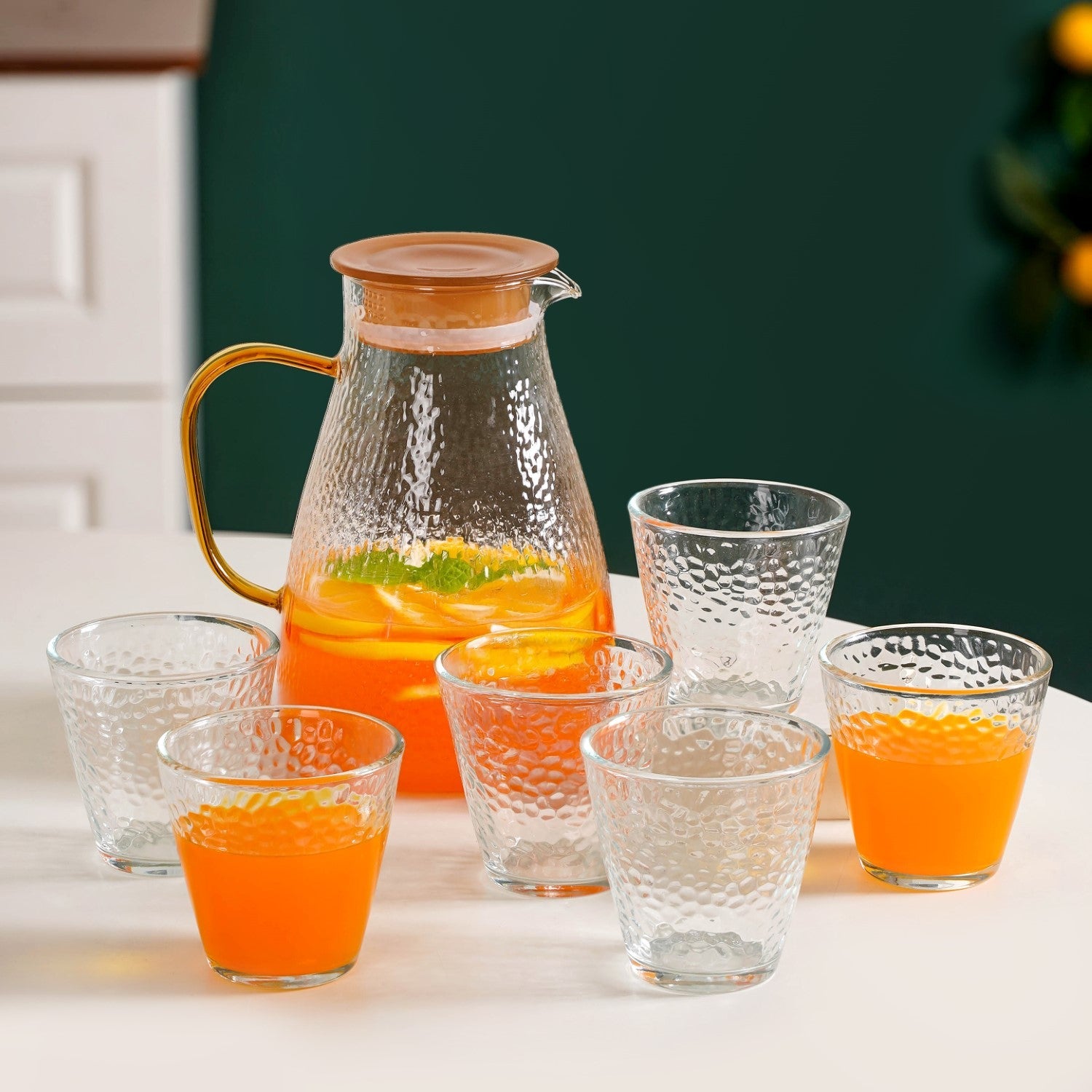 Clear glass pitcher and cups filled with orange juice on a white surface.